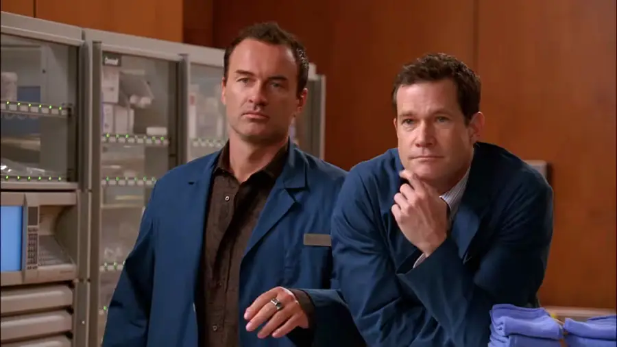 Two men in blue lab coats stand in a medical storage room. One man leans casually on a counter, while the other stands beside him, both with neutral expressions. Medical supplies line the shelves in the background.