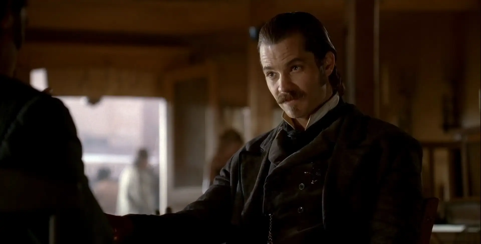 A man with slicked-back hair and a mustache, dressed in dark old-fashioned clothing, sits indoors at a table, looking intently at someone off-screen. The background is softly lit.