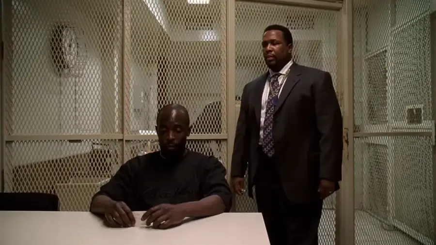 A man sits at a white table in an interrogation room, looking down, while another man in a suit stands behind him. A metal mesh divides the room.