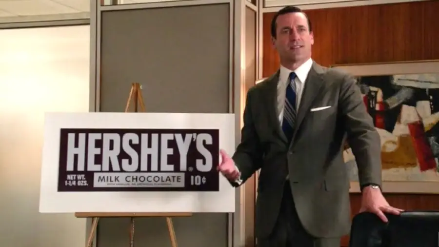 A man in a suit stands next to a large Hershey’s Milk Chocolate bar sign on an easel in an office setting, gesturing as if giving a presentation.