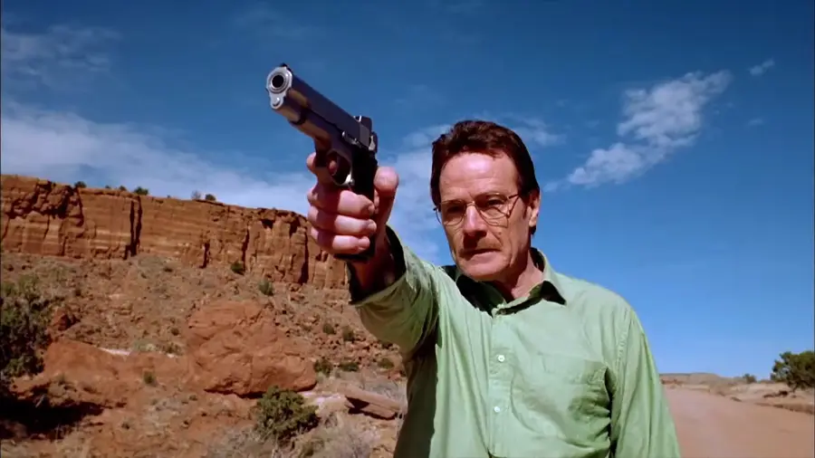A man in a green shirt stands on a dirt road in a desert landscape, pointing a handgun directly toward the camera under a blue sky with scattered clouds.