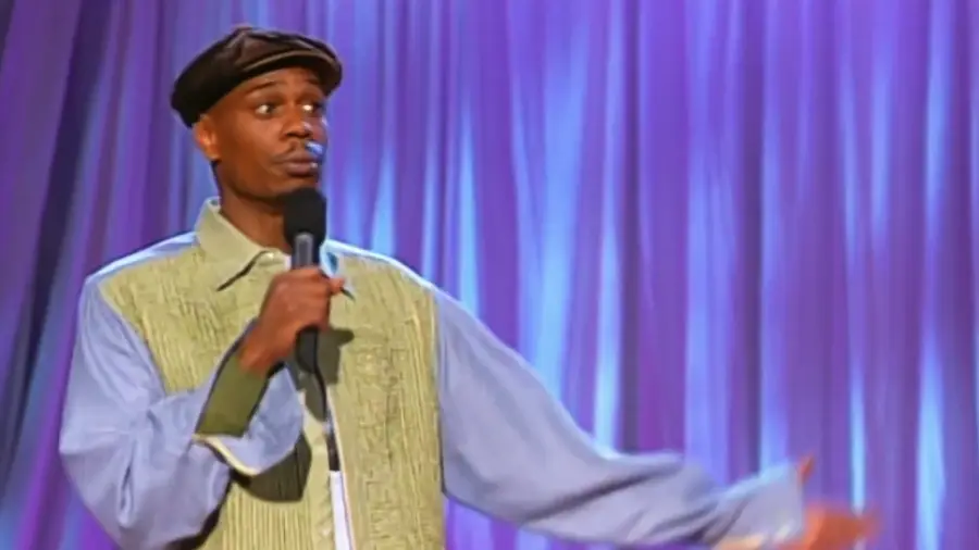 A comedian in a cap and patterned shirt holds a microphone, gesturing with one hand, performing on stage with a purple curtain backdrop.