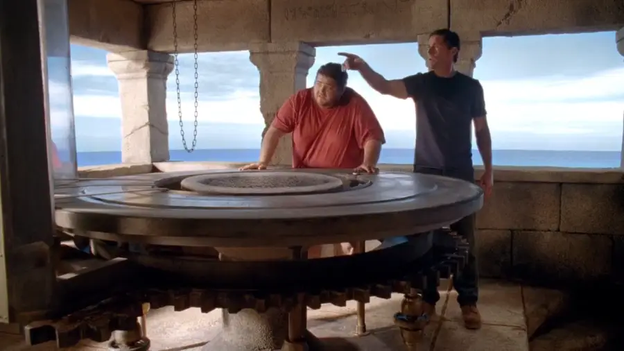 Two men stand in a stone room with ocean views. One man touches a large, round stone wheel in the center, while the other reaches over to pat his head. Sunlight streams through arched windows.