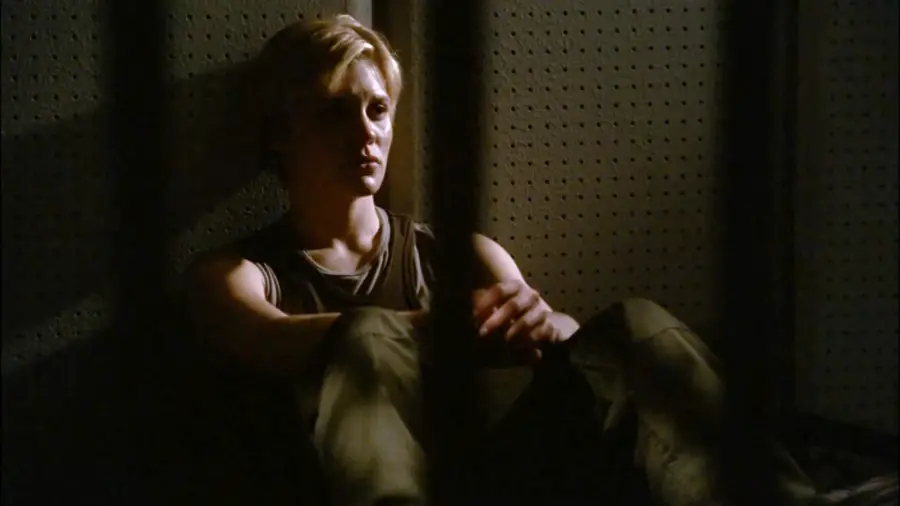 A person with short blonde hair sits on the floor in a dimly lit room, leaning against a wall behind metal bars, looking thoughtful or distressed.