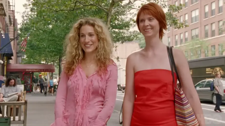 Two women walk down a city sidewalk smiling. One wears a pink blouse, the other a red dress and striped bag. Trees, buildings, and outdoor seating are in the background.