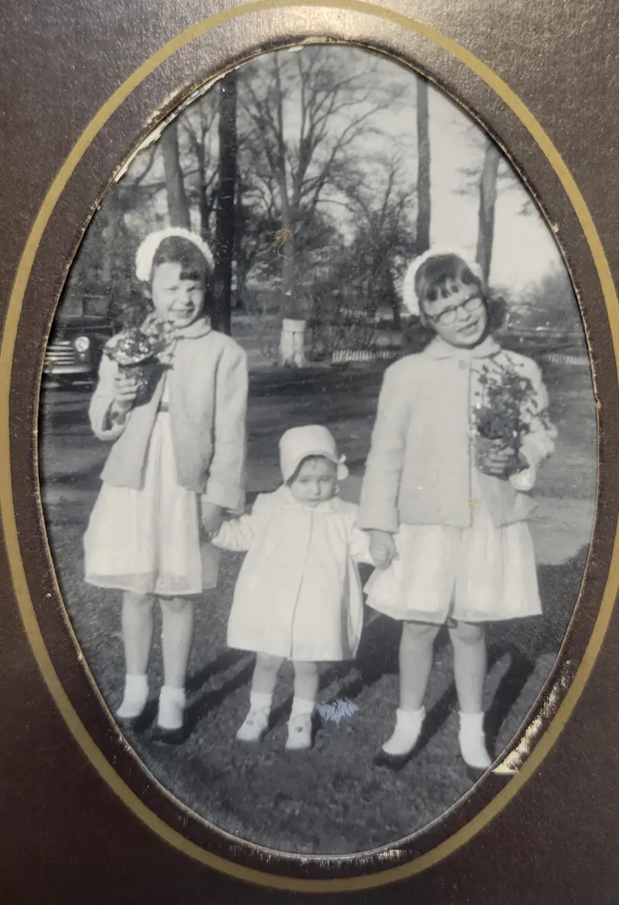 Three young girls in light-colored coats and dresses stand outside, holding hands. The two older girls hold bouquets, and all three wear hats. The photo is black and white with an oval frame. Trees and a car are in the background.