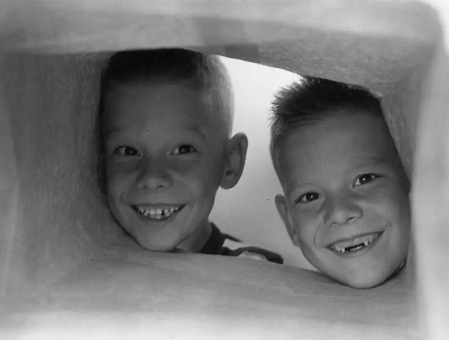 Two young boys with short hair smile through an opening in a soft, light-colored structure, showing their teeth with gaps. The lighting is bright, highlighting their happy expressions.