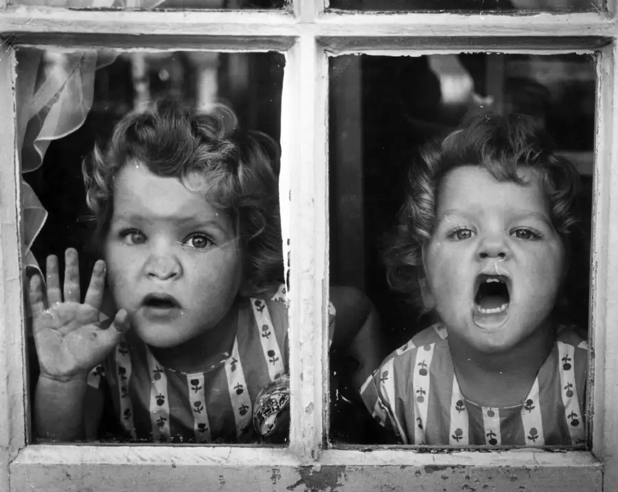 Two young children with curly hair and striped shirts press their faces against a windowpane. One has a hand raised to the glass, while the other has their mouth open wide as if shouting or calling out.