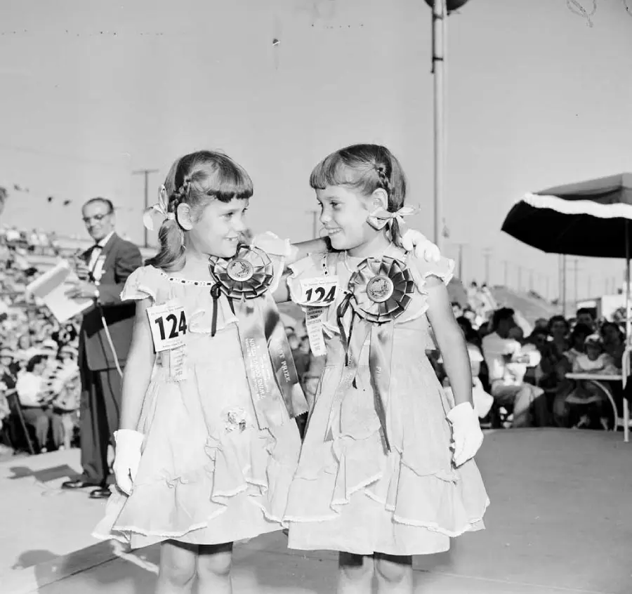 Two young girls in matching dresses, gloves, and large award ribbons stand on a stage, smiling at each other. They wear number 124 and appear to be in a competition; a crowd and judge are visible in the background.