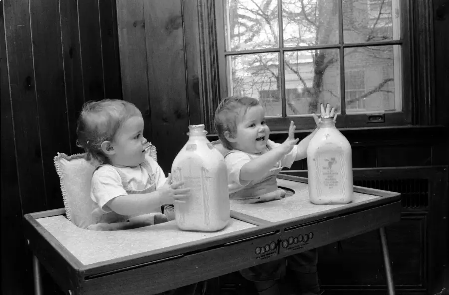 Two toddlers sit side by side in high chairs by a window, each with a large glass bottle of milk. One child smiles and reaches out, while the other looks to the side, holding the bottle.