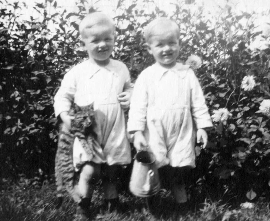 Two young children in similar outfits stand outdoors. One holds a striped cat, and the other holds a tin pail. Tall plants and flowers fill the background.