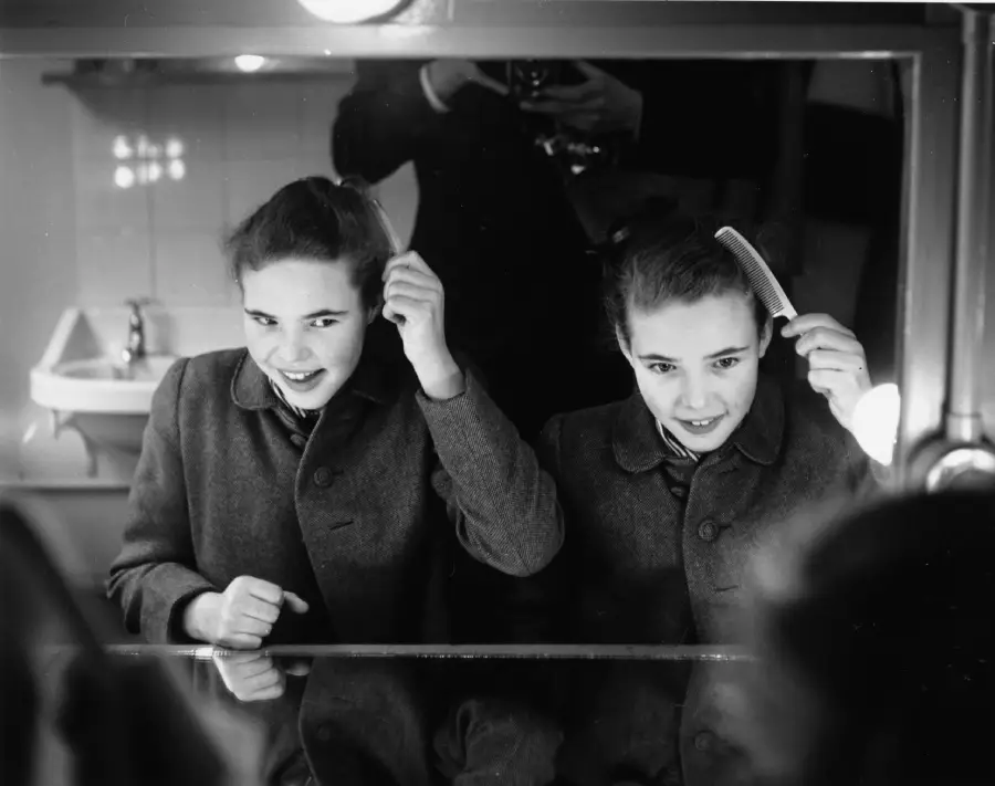 Two girls stand side by side in front of a mirror, brushing and arranging their hair, with a sink in the background and a person reflected, holding a camera.