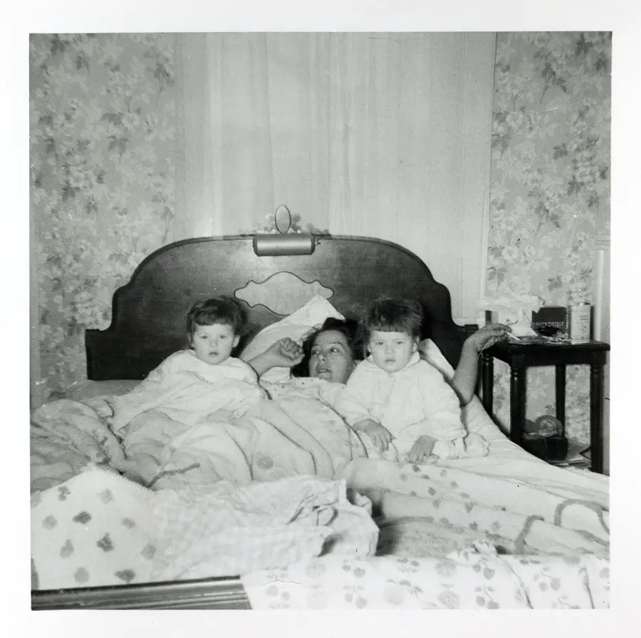 A woman lies in bed between two young children. They are all covered with blankets, and the room has floral wallpaper. A nightstand with various items is beside the bed.