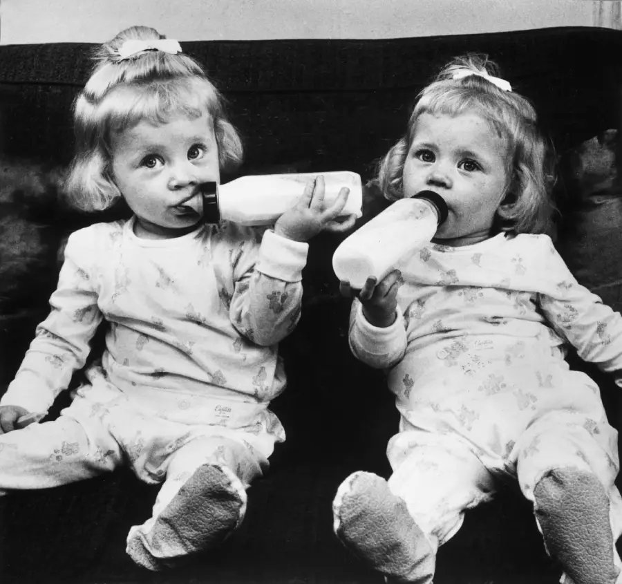 Two young twin girls sit on a couch, each wearing matching pajamas and drinking from baby bottles. They have similar hairstyles with bows and are looking toward the camera.