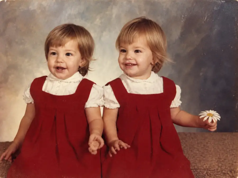 Two young children with short light brown hair wear matching red dresses and white shirts. They are sitting together; one smiles and holds a daisy. The background is a soft, blurred studio portrait backdrop.