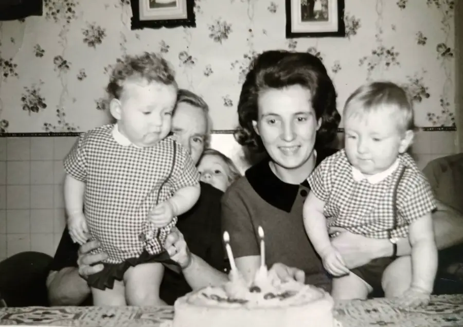 A black-and-white photo of a woman holding two toddlers in matching outfits, with a man and another child partially visible behind them. A cake with two candles is on the table in front.