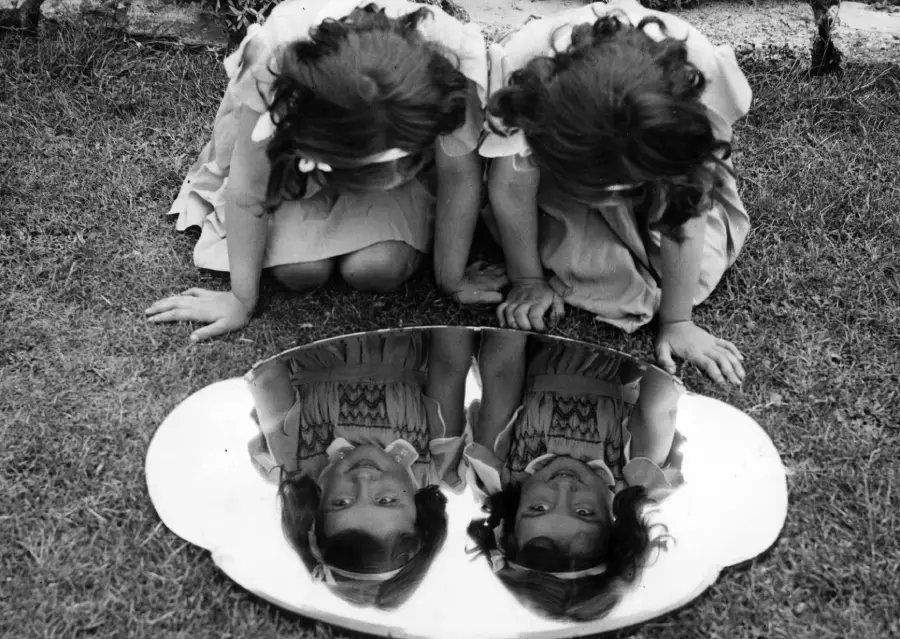 Two young girls in dresses kneel on grass, looking down at their reflections in an oval mirror placed on the ground, creating the illusion of four children.
