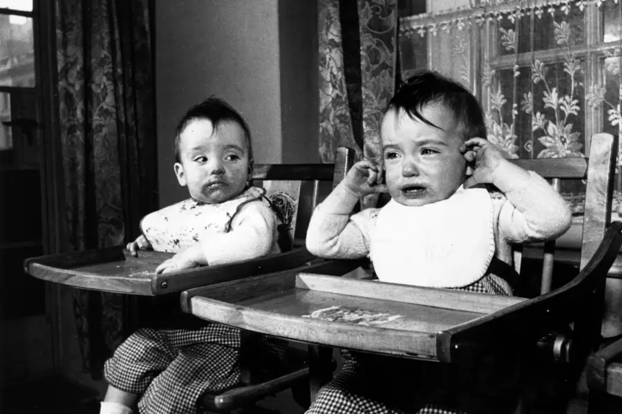 Two toddlers sit in wooden high chairs. One child is calm, looking to the side, while the other is crying and rubbing their ears. Both wear bibs and have food smeared on their faces. A window with lace curtains is behind them.