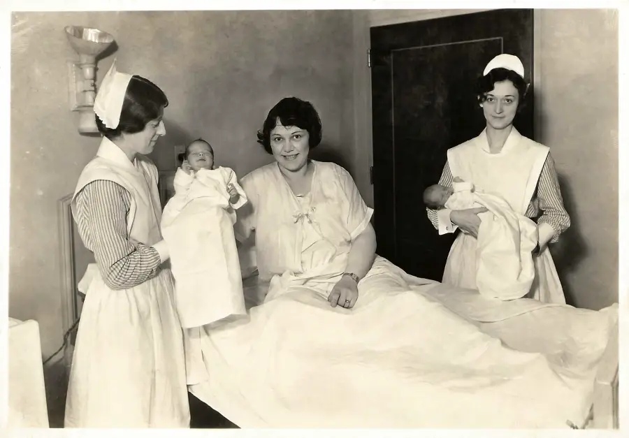 A woman sits on a hospital bed holding a newborn, with two nurses in uniforms nearby, one holding another baby. The scene appears to be from the early 20th century.