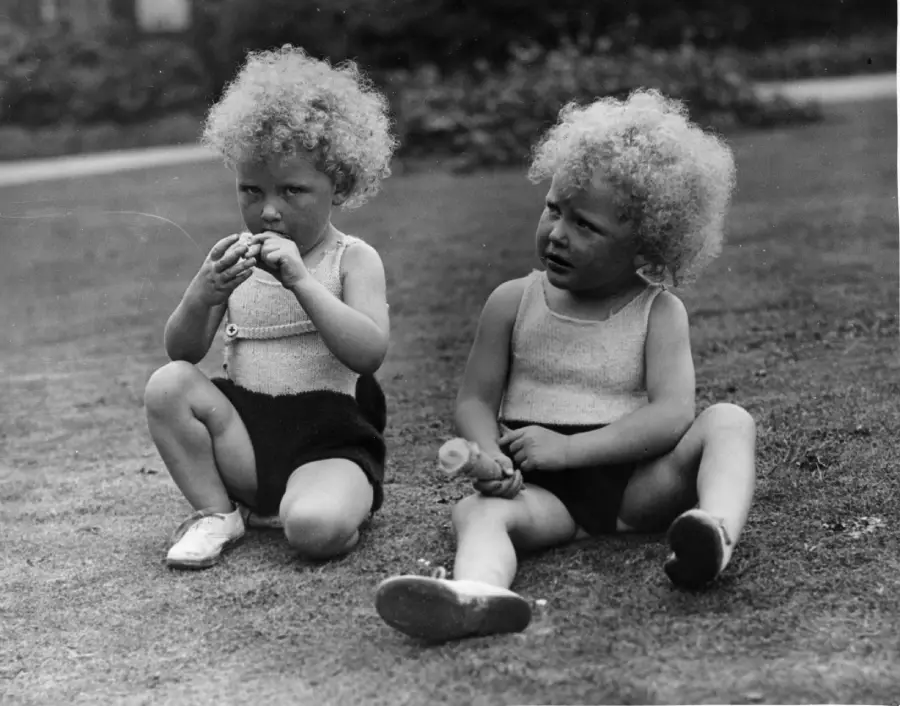Two young children with curly blond hair sit on the grass. Both wear light sleeveless tops and dark shorts. One child is eating, while the other looks away, holding food. The background is blurry and outdoors.