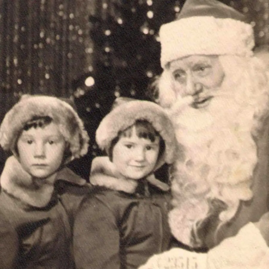 Two young children in winter coats and fur hats pose with a man dressed as Santa Claus. The background has a decorated Christmas tree and sparkling lights, suggesting a festive holiday setting. The image is in black and white.