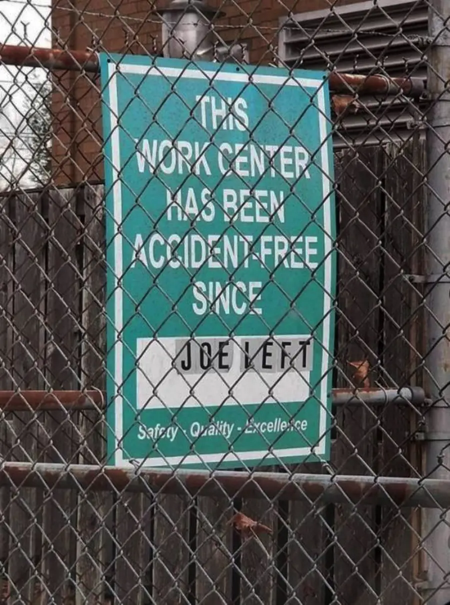 A work safety sign reads, “This work center has been accident-free since JOE YETT,” showing no actual date. The sign hangs on a chain-link fence outside an industrial area.