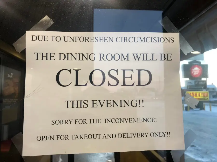 A printed sign taped to a window reads, “Due to unforseen circumcisions, the dining room will be closed this evening!! Sorry for the inconvenience! Open for takeout and delivery only!!”