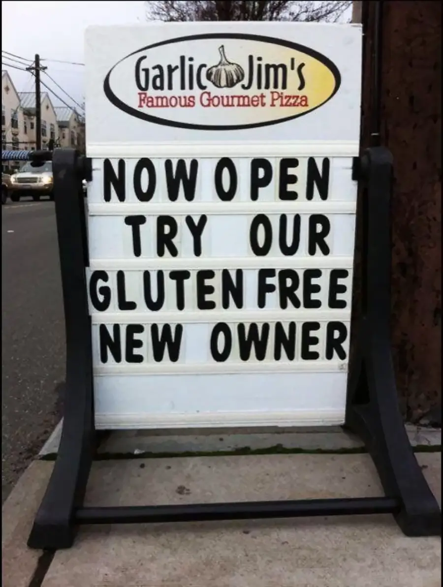 A sidewalk sign reads: “Garlic Jim’s Famous Gourmet Pizza. NOW OPEN TRY OUR GLUTEN FREE NEW OWNER.” The message appears unintentionally humorous.
