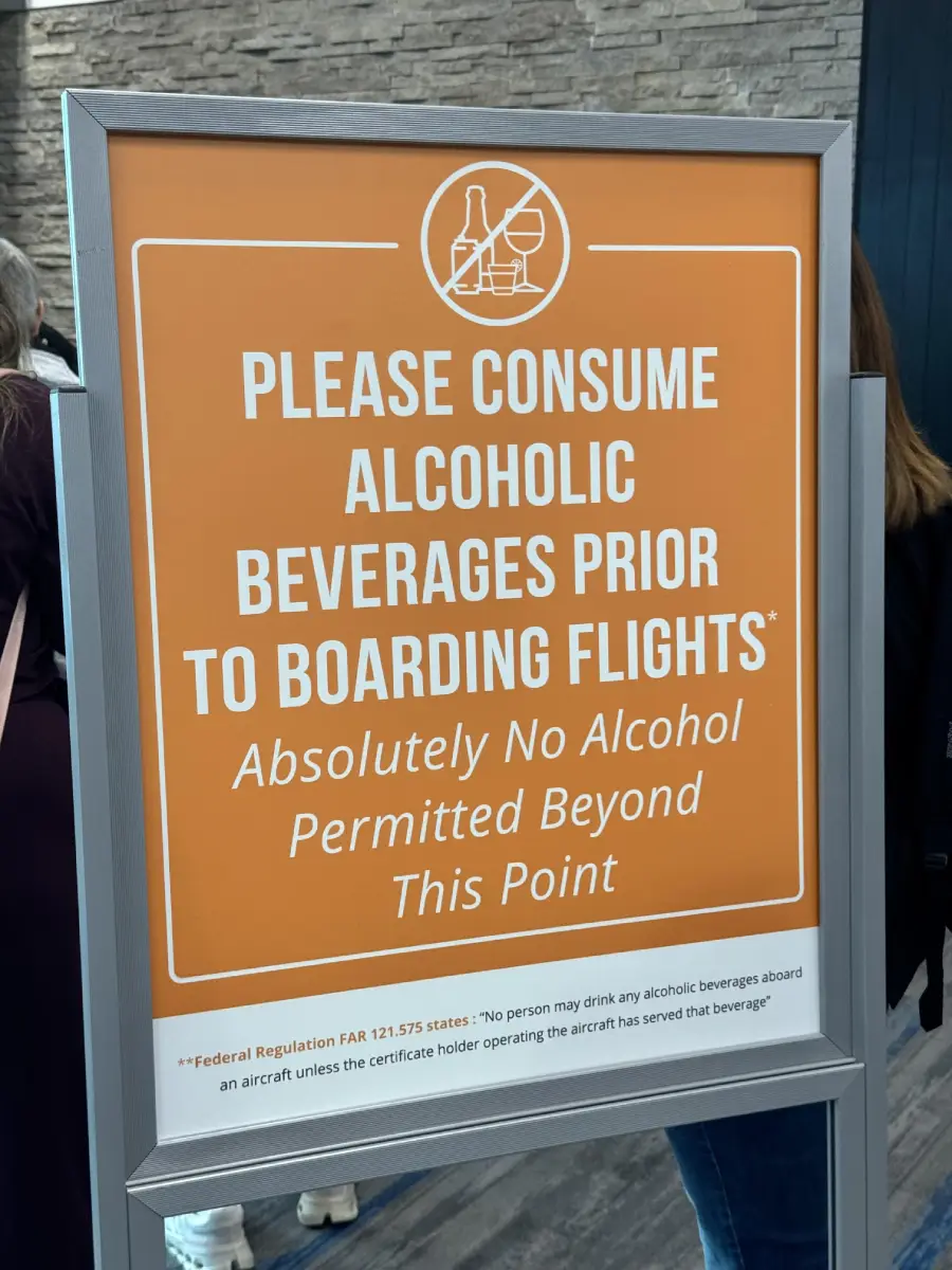 An orange sign tells passengers to finish alcoholic beverages before boarding flights, stating no alcohol is allowed past this point. People stand nearby.