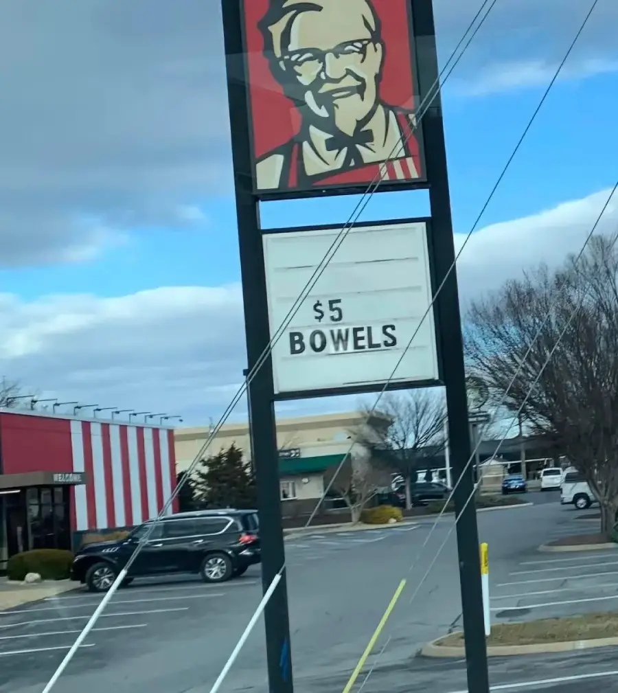A KFC sign displays “$5 BOWELS” instead of “bowls.” The parking lot and a few cars are visible, with a cloudy sky in the background.
