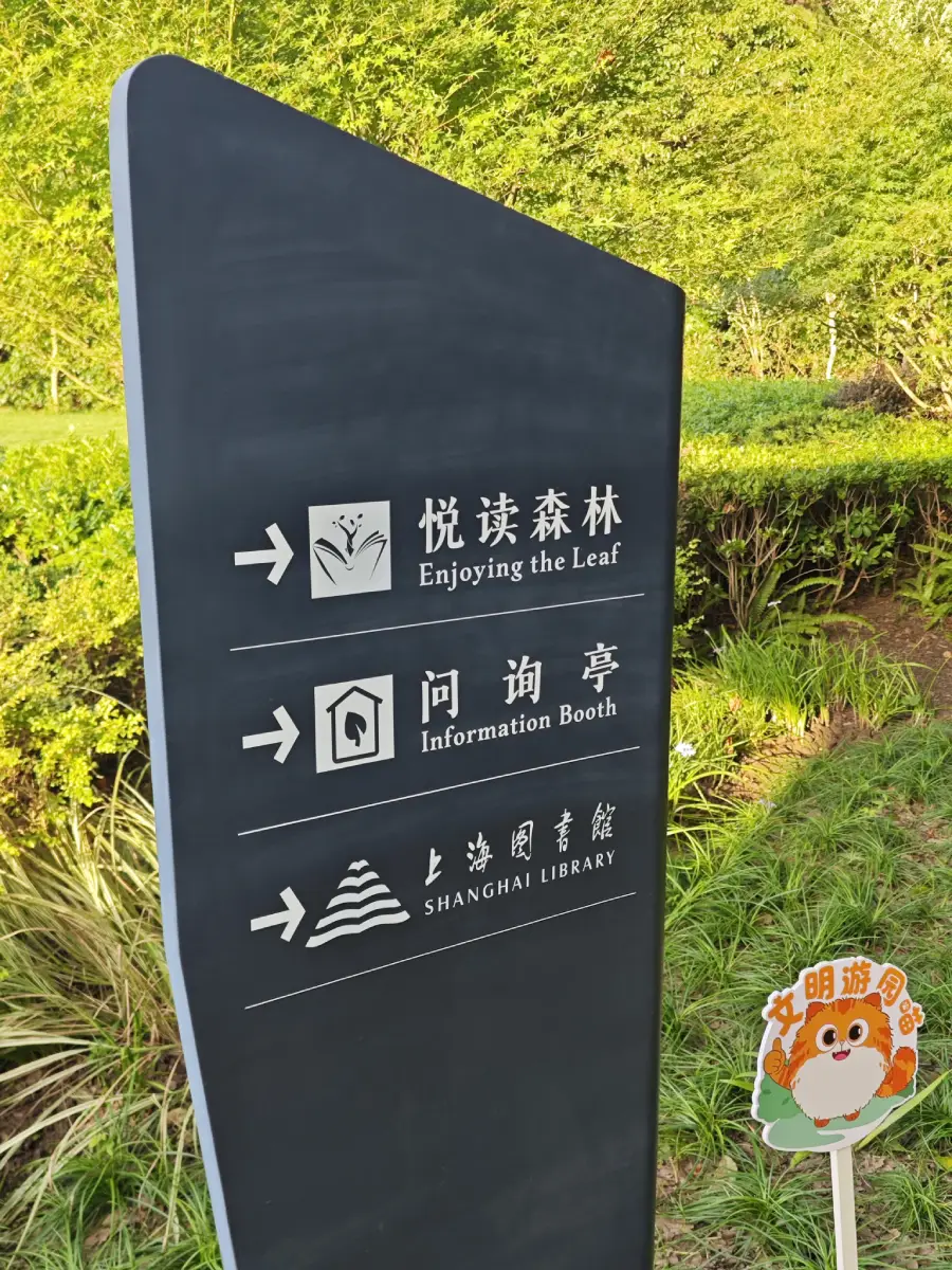 A black sign with white text and arrows points to “Enjoying the Leaf,” “Information Booth,” and “Shanghai Library,” in both English and Chinese, amid green foliage. A small cartoon animal sign is in the bottom right.