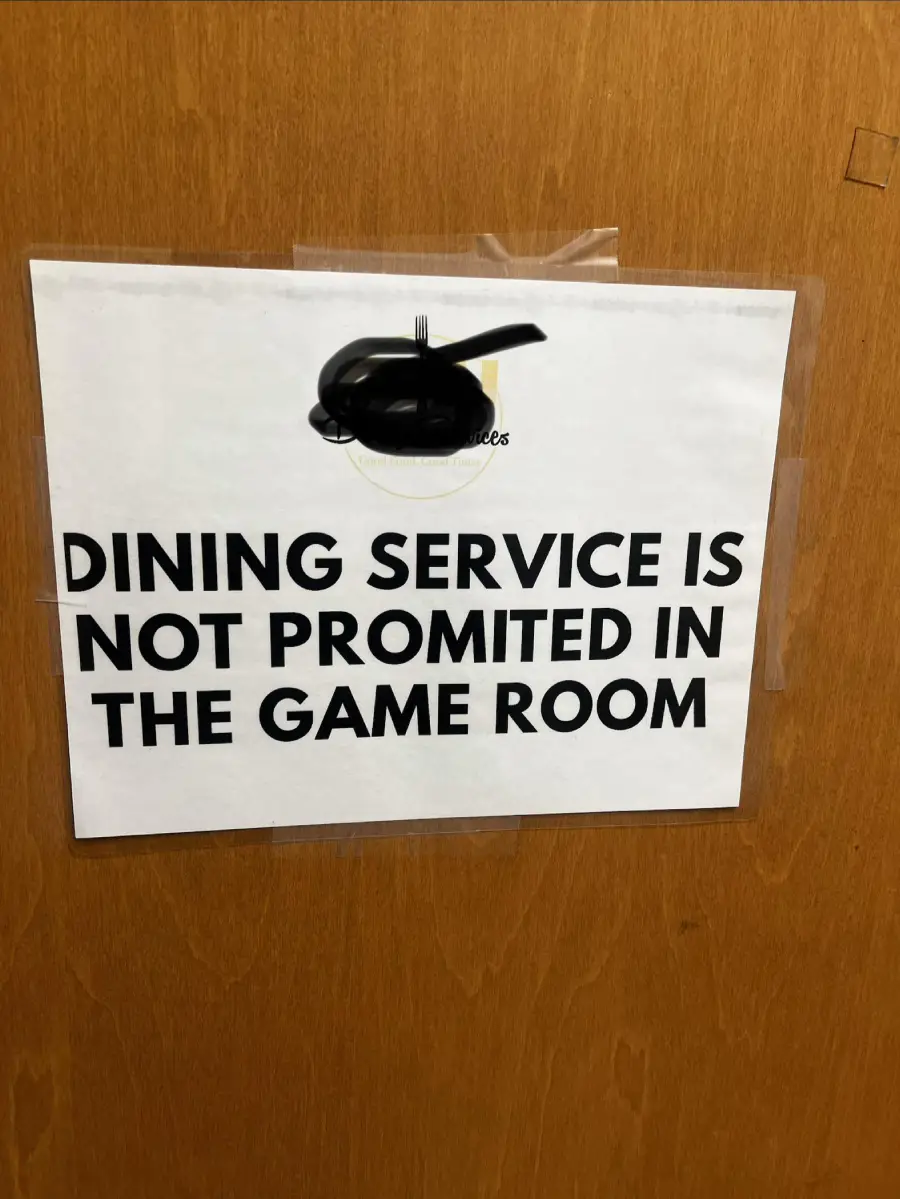 A printed sign taped to a wooden door reads, “DINING SERVICE IS NOT PROMITED IN THE GAME ROOM.” The word “promited” is a misspelling of “permitted.” A black headset is taped above the sign.