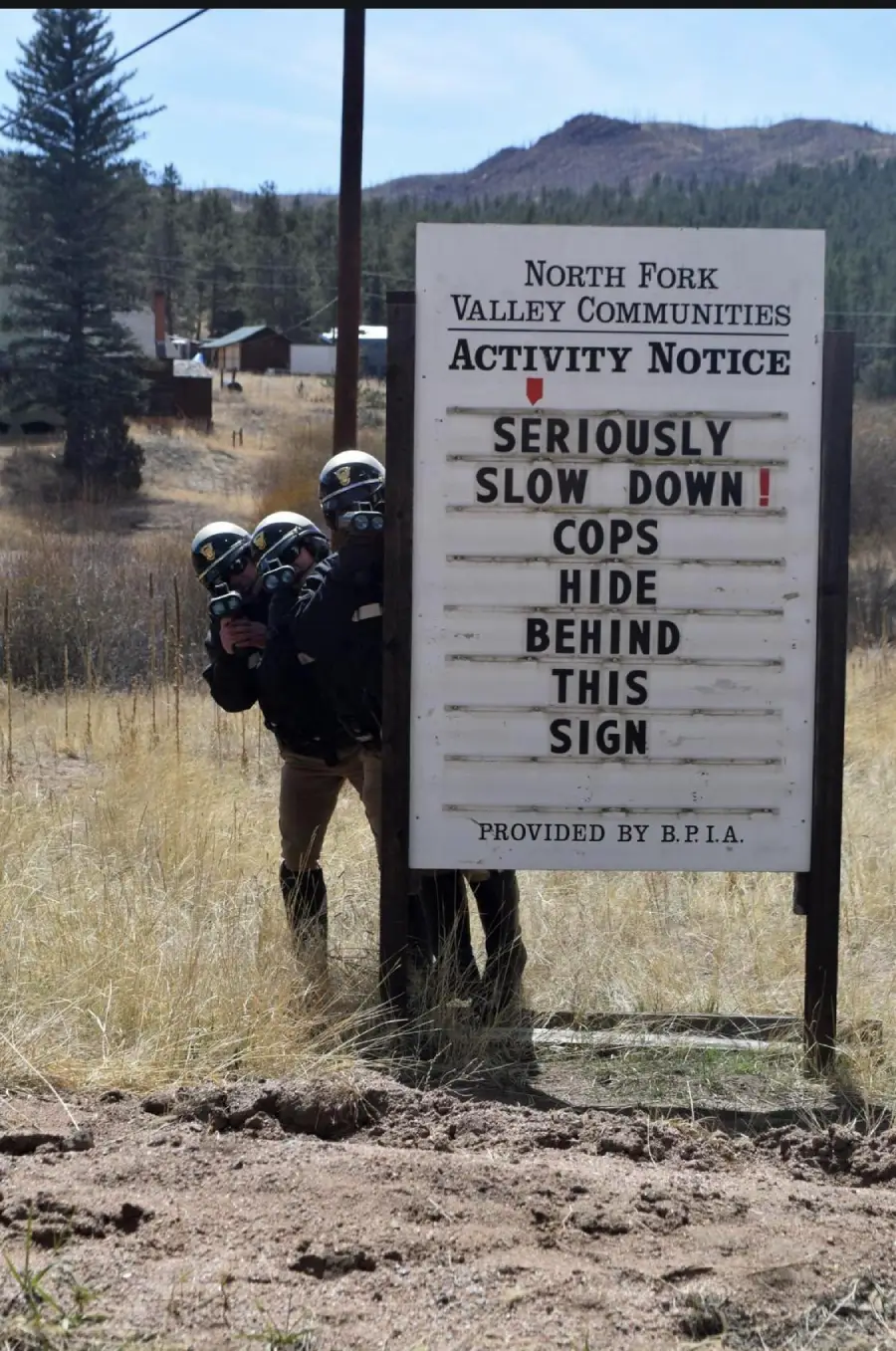 Two police officers in helmets and gear stand behind a community activity sign. The sign reads: “SERIOUSLY SLOW DOWN! COPS HIDE BEHIND THIS SIGN.” The officers are holding radar guns, pointing them towards the road. Rural landscape in the background.