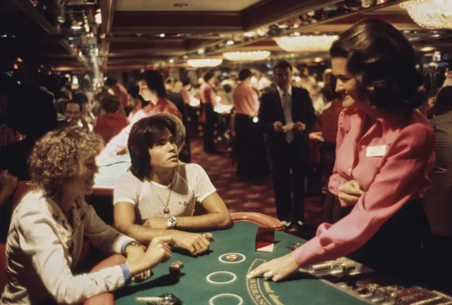Two people sit at a casino table with cards and chips, while a woman in a pink blouse stands nearby talking to them. The casino is crowded, with people and lights in the background.