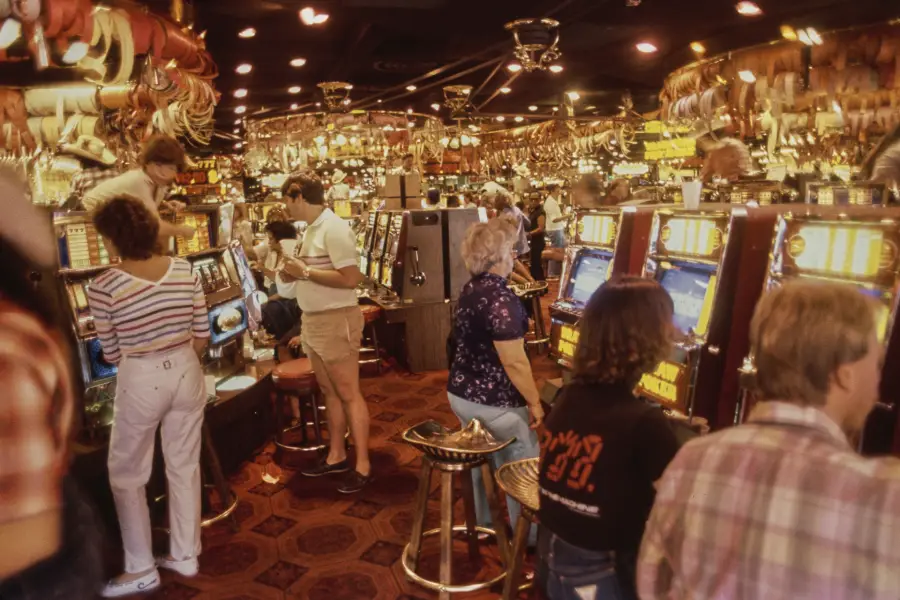People playing slot machines in a crowded, brightly lit casino with a patterned carpet and reflective ceiling.