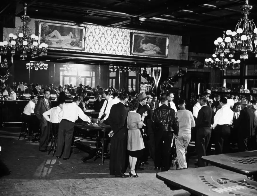 Black and white photo of a crowded casino with people gathered around gaming tables, chandeliers hanging from the ceiling, and paintings of reclining figures on the walls in the background.