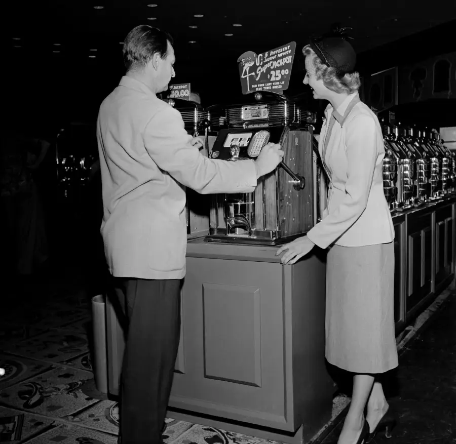 A man and woman stand at a vintage slot machine. The man pulls the lever while the woman watches and smiles. Other slot machines are visible in the background. The scene appears to be from the mid-20th century.