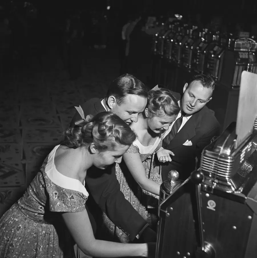 Four people in formal attire, two men and two women, are closely gathered around slot machines in a dimly lit room, appearing excited as they play.