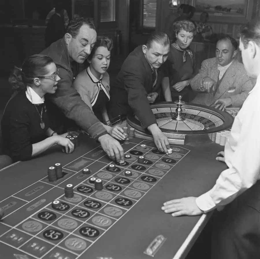 A group of people gathered around a roulette table in a casino, placing bets with chips as a croupier oversees the game.