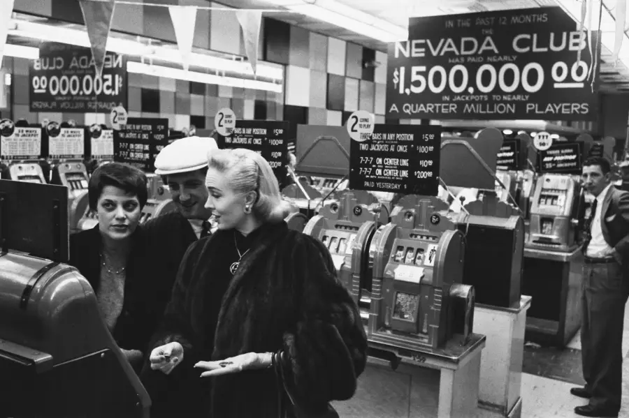 Three people stand near slot machines inside a casino. Large signs advertise $1,500,000 in jackpots at the Nevada Club. The environment is busy, with rows of machines and other people in the background.