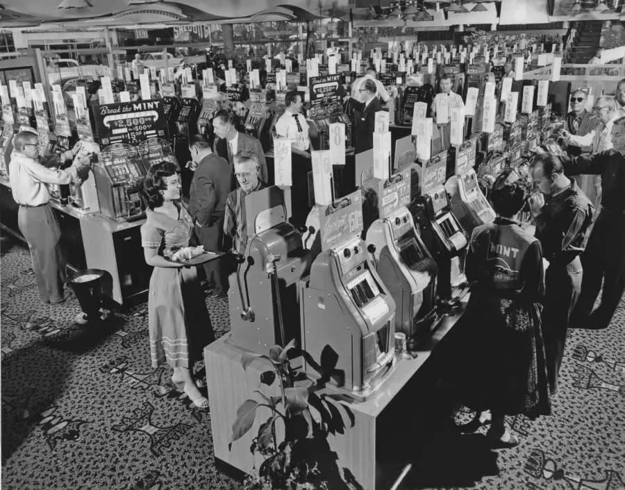 Black and white photo of people playing rows of slot machines in a busy casino. Many are standing and pulling levers, while others watch. The scene appears lively and vintage, likely from the mid-20th century.