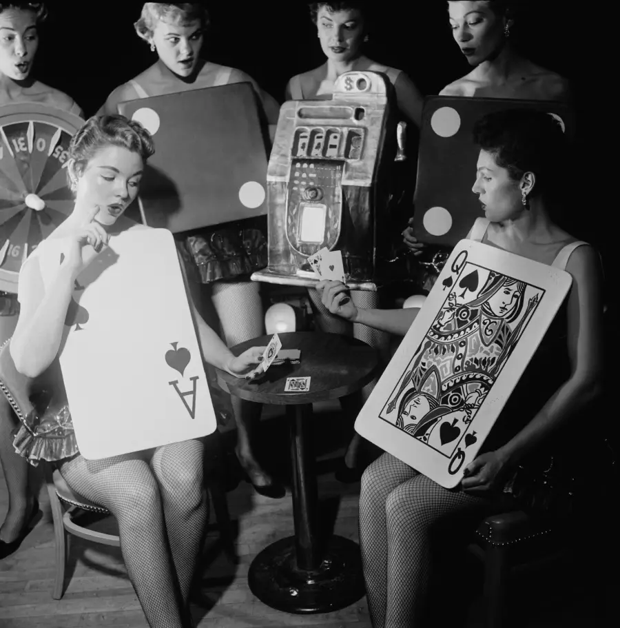 Five women in costumes resemble casino games, including cards, a roulette wheel, dice, and a slot machine, as they sit and play cards around a small table, appearing lively and engaged.