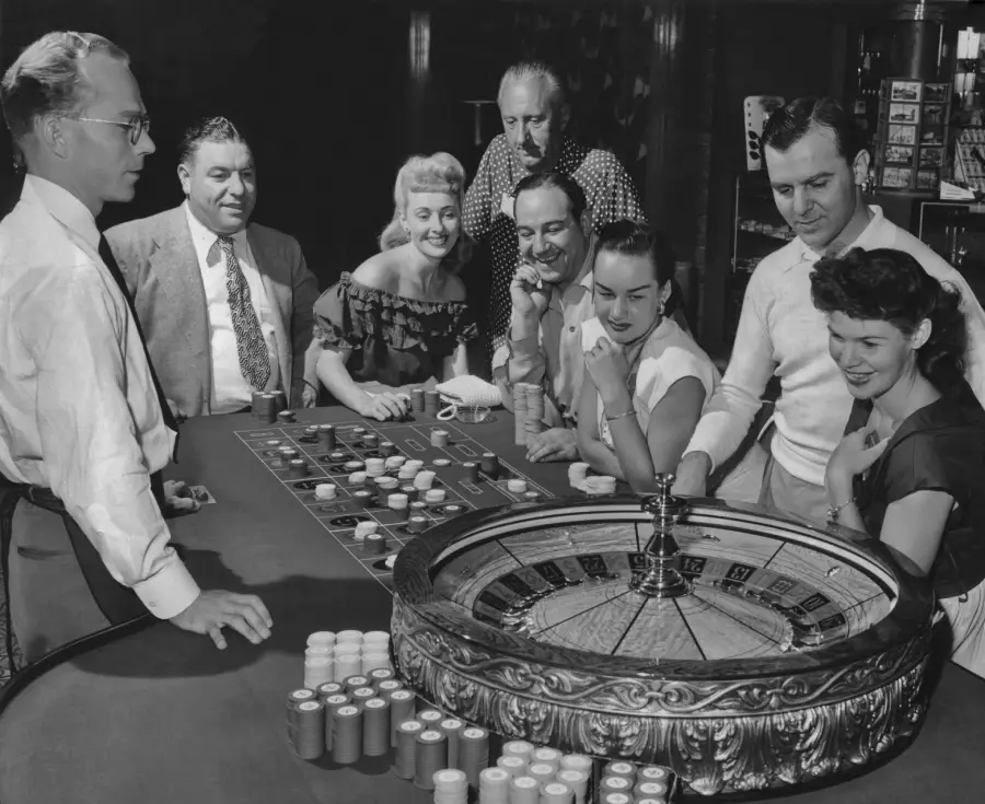 A group of well-dressed people gathers around a roulette table in a casino, watching the spinning wheel and awaiting the result, with chips and a croupier in view.