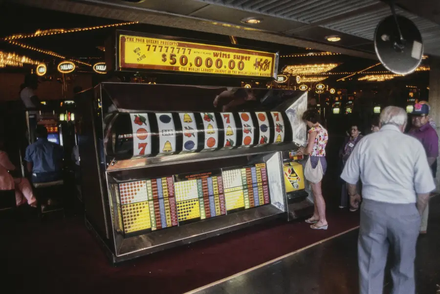 People stand near a giant slot machine in a casino, with colorful lights and other slot machines in the background. A sign above advertises a $50,000 prize. The scene captures a lively, busy casino atmosphere.