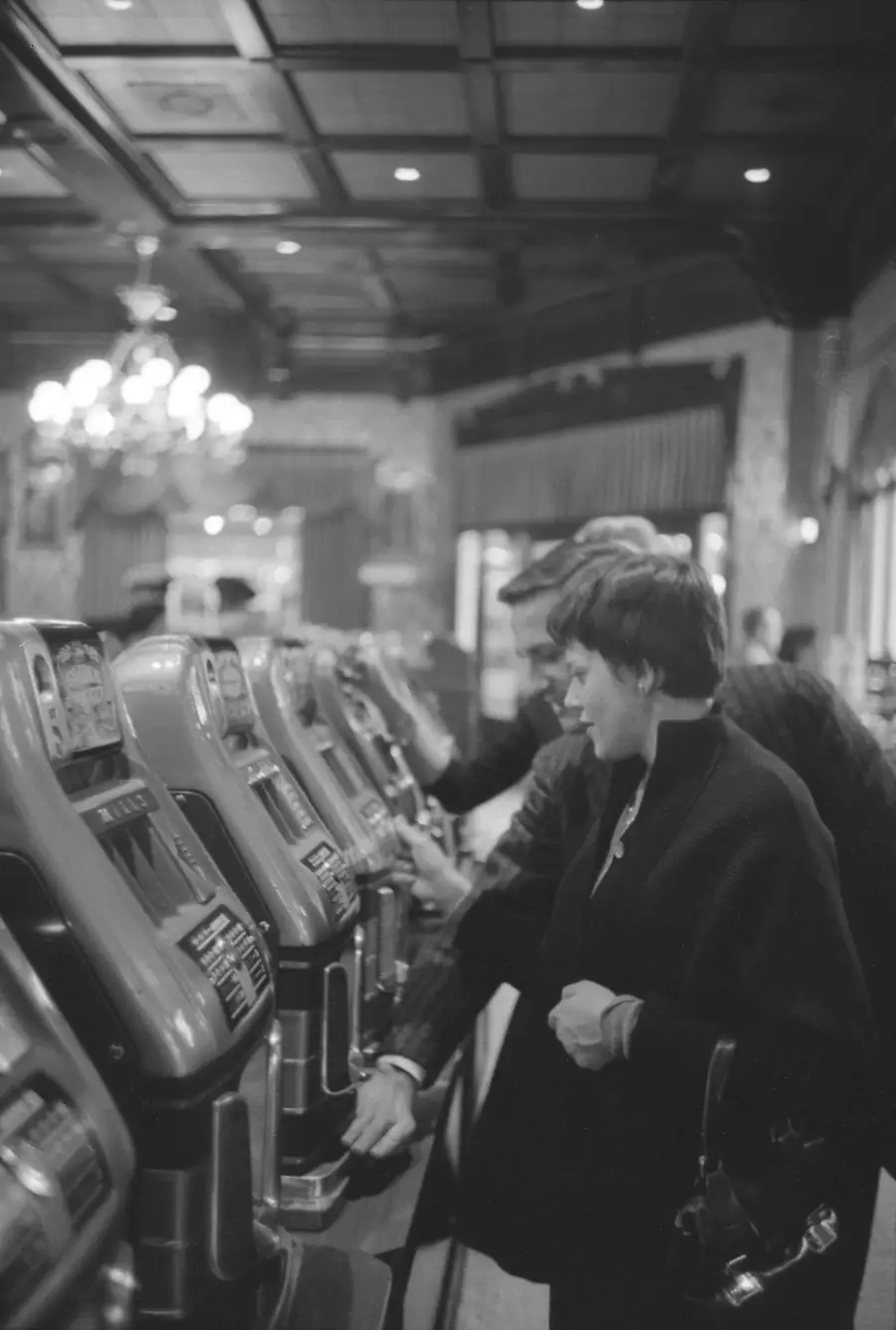 A woman and man stand side by side playing slot machines in a vintage casino, with chandeliers and wood-paneled ceiling visible in the background. Black and white photo.