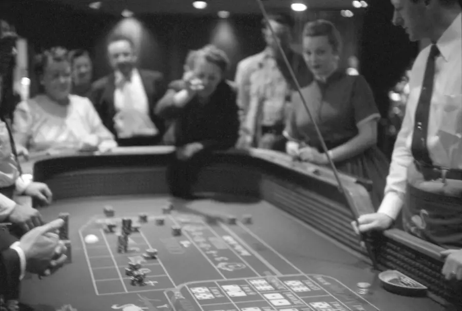 A group of people gather around a casino craps table, watching intently as one person throws dice. Chips and game markings are visible on the table. The scene is lively and dimly lit.