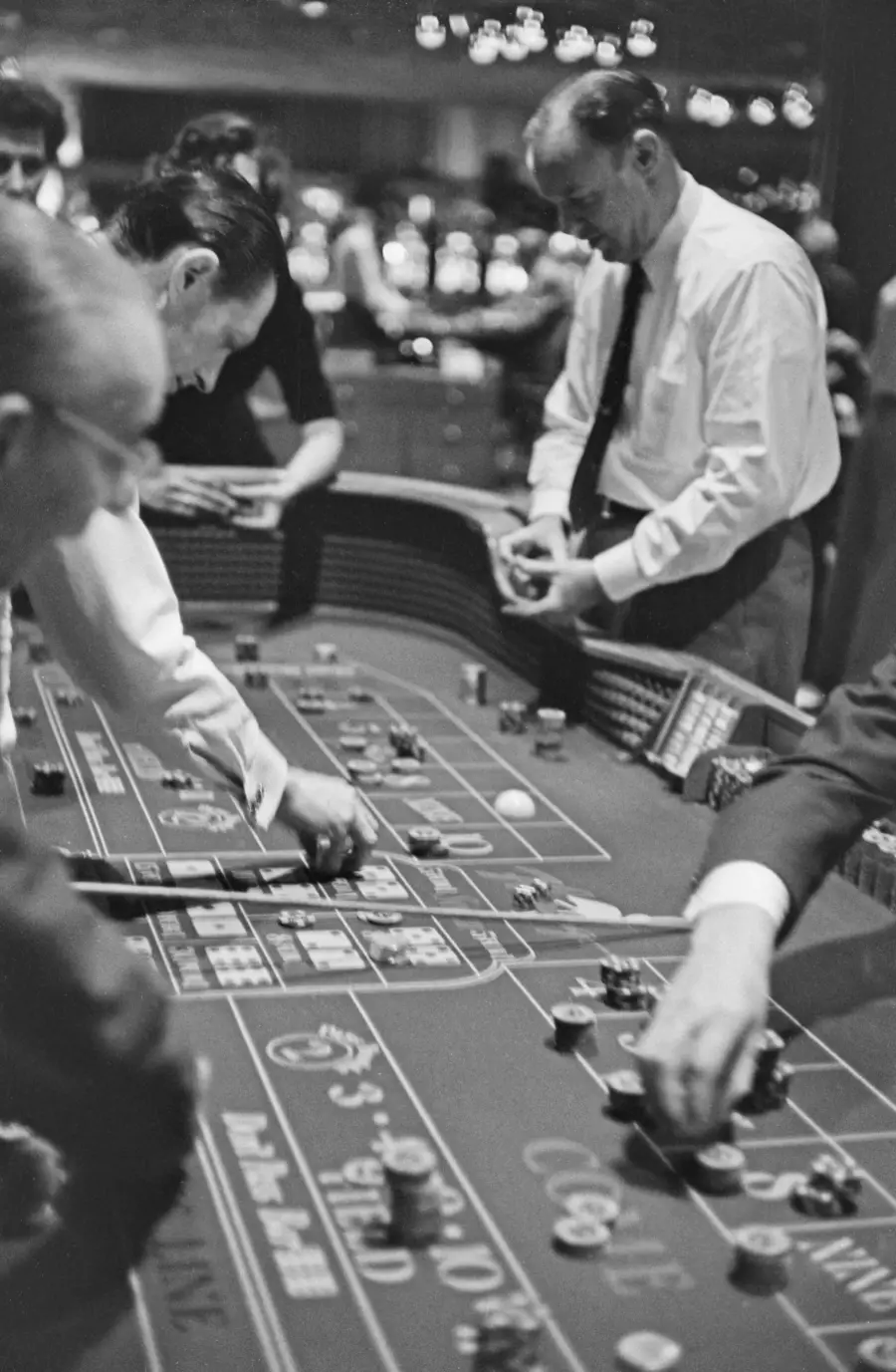 Black and white photo of people standing around a casino craps table, placing bets and throwing dice, with chips and cards on the table. Background shows a busy, lively atmosphere with blurred lights.