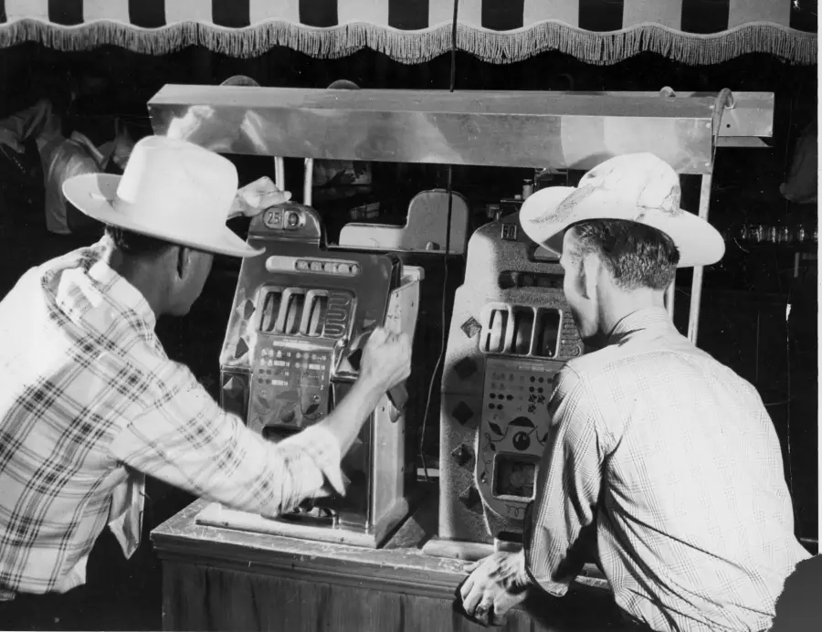 Two men in cowboy hats and plaid shirts play vintage slot machines side by side under a striped canopy.