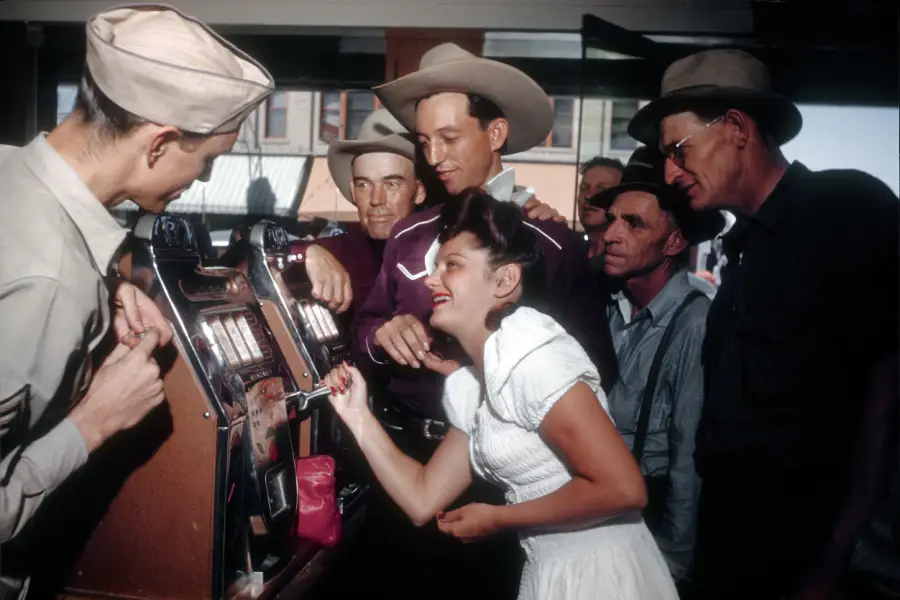 A woman in a white dress plays a slot machine, while several men in hats and uniforms watch her and smile in a lively, vintage scene.