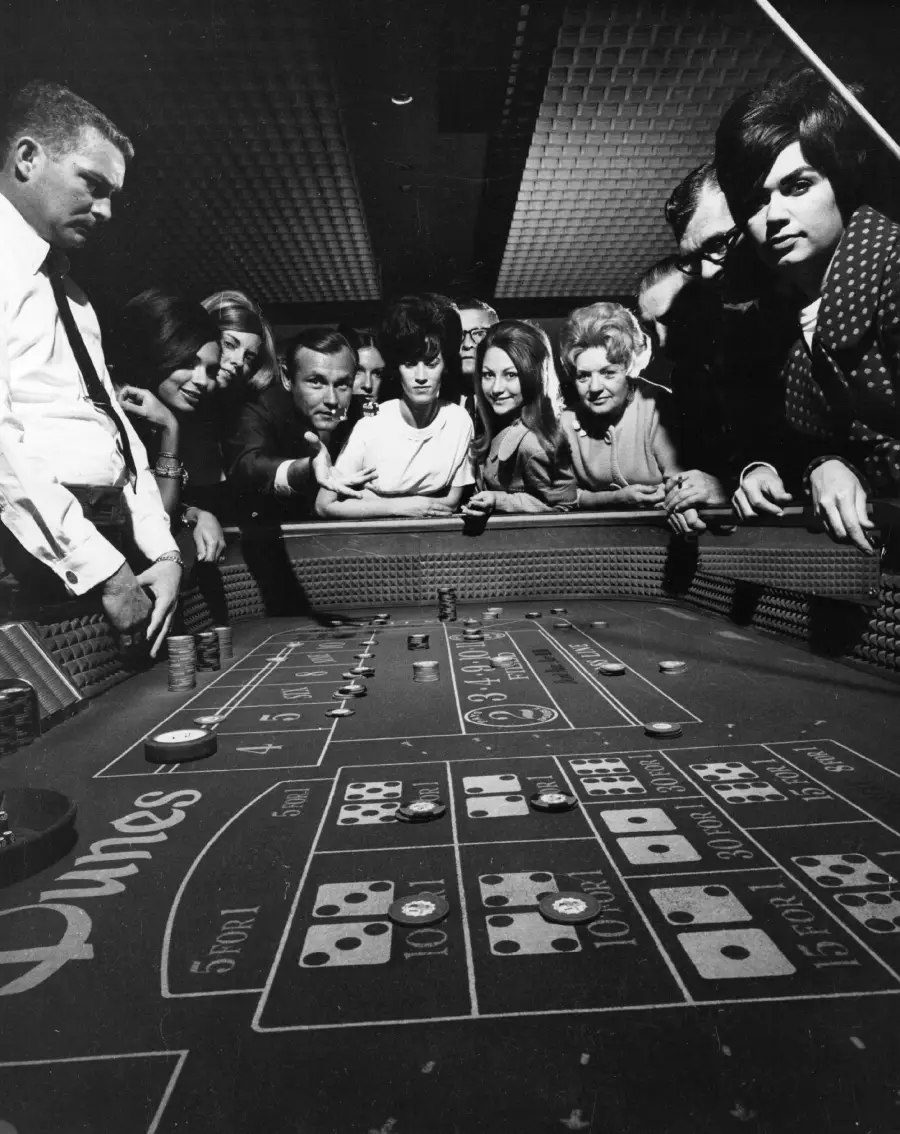 A group of people, men and women, closely watch a craps table in a casino, appearing tense and focused as dice are rolled. Chips and betting areas are visible in the foreground.